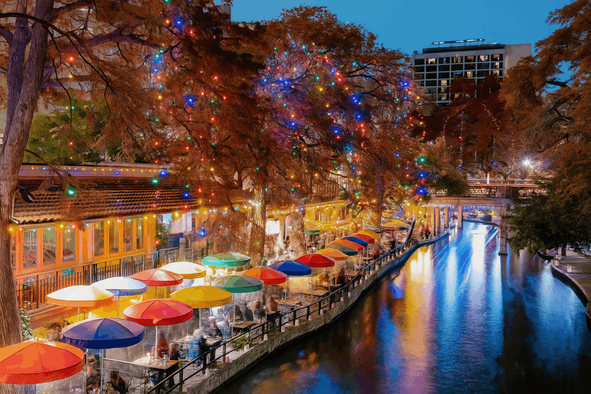 The San Antonio River Walk at night decorated with colorful holiday lights, featuring glowing trees and vibrant umbrellas reflecting on the water.