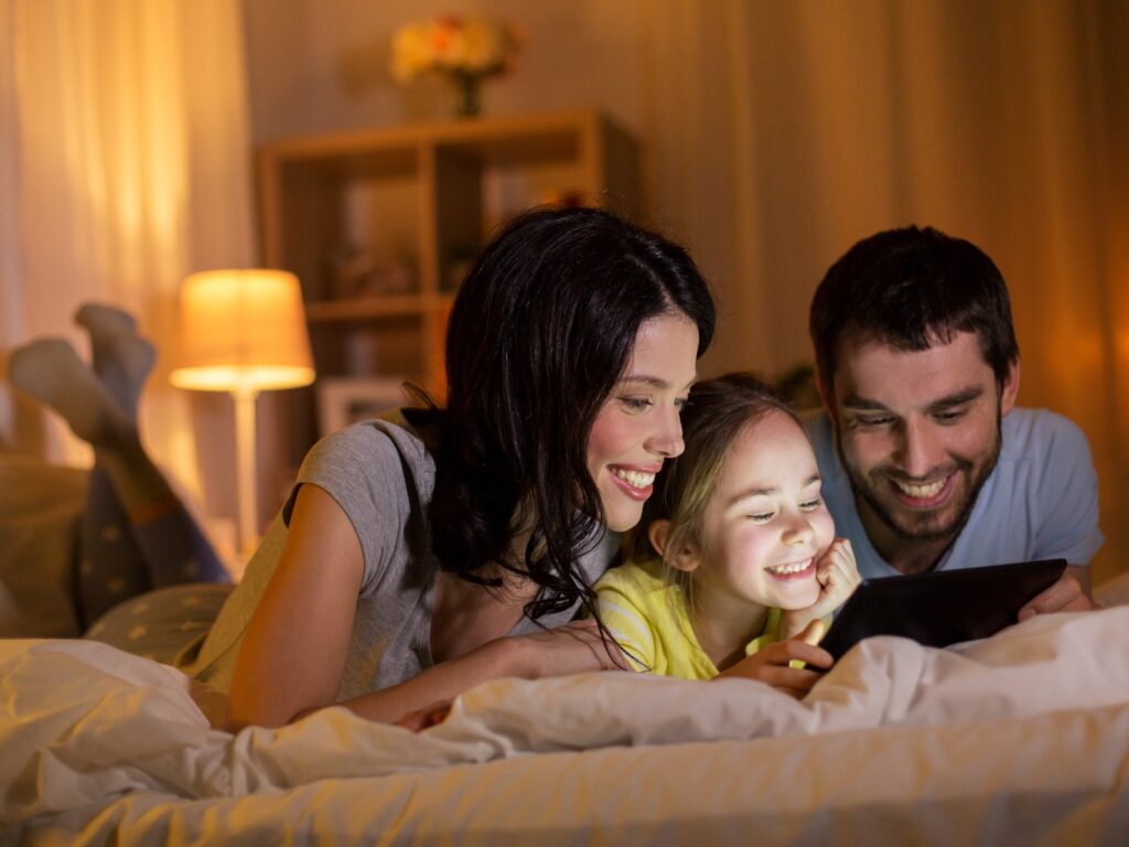 Happy mother, father and little daughter with tablet in bed at night at home