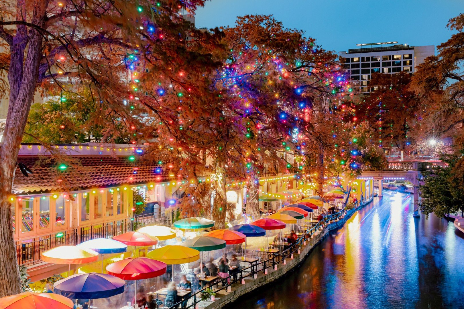 San Antonio River Walk at night during the holidays, featuring colorful umbrellas, festive Christmas lights, and reflections on the water at popular restaurants and shops.